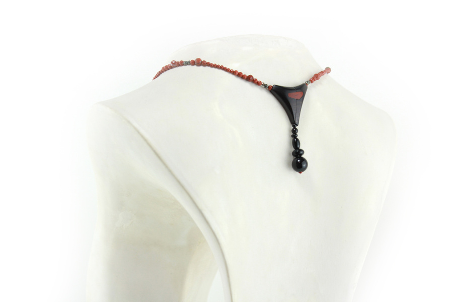 Design-Choker aus exotischen Hölzern und rotem Jaspis, mit einer Ranke aus schwarzem Ebenholz als Anhänger, die mit einem Kreis aus kleinen, glitzernden, goldbronzenen Pyritperlen befestigt ist, verbunden mit einem gebogenen Schaukelstück aus dunkel cremefarbenem, holzigem Olivenholz, und einer Perlenschnur aus ziegelroten, mit zwei Pyritperlen punktierten Perlen, die von beiden Seiten des Stücks zu einem dreieckigen, magnetischen Puzzle-Verschluss mit einem Anhänger aus schwarzen Perlen als Verschluss führt.