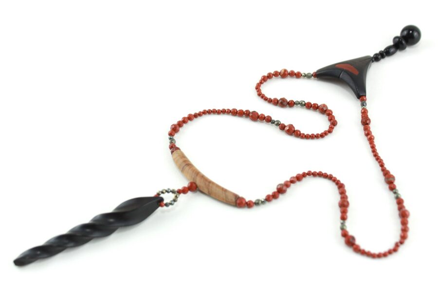 Choker design en bois exotiques & Jaspe rouge, avec pour pendentif une vrille en bois d'ébène noir, attachée par un cercle de petite perle de pyrité bronze doré scintillant, reliée à une pièce balancelle courbée en bois d'olivier crème foncé boisé, et un fil de perles rouge brique ponctués de deux de pyrite partent des deux côté de la pièce jusqu'au fermoir puzzle aimanté de forme triangulaire, avec un pendant de perles noires en fermeture