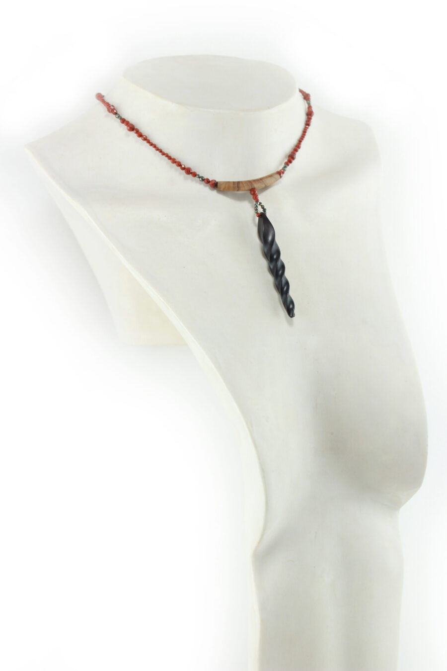 Choker design en bois exotiques & Jaspe rouge, avec pour pendentif une vrille en bois d'ébène noir, attachée par un cercle de petite perle de pyrité bronze doré scintillant, reliée à une pièce balancelle courbée en bois d'olivier crème foncé boisé, et un fil de perles rouge brique ponctués de deux de pyrite partent des deux côté de la pièce jusqu'au fermoir puzzle aimanté de forme triangulaire, avec un pendant de perles noires en fermeture