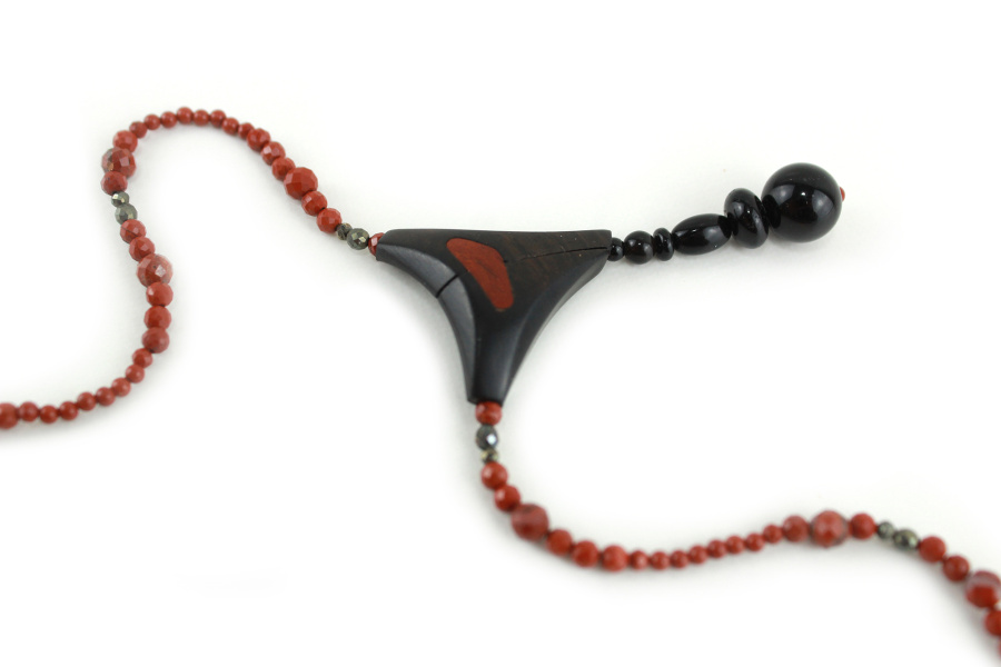 Design-Choker aus exotischen Hölzern und rotem Jaspis, mit einer Ranke aus schwarzem Ebenholz als Anhänger, die mit einem Kreis aus kleinen, glitzernden, goldbronzenen Pyritperlen befestigt ist, verbunden mit einem gebogenen Schaukelstück aus dunkel cremefarbenem, holzigem Olivenholz, und einer Perlenschnur aus ziegelroten, mit zwei Pyritperlen punktierten Perlen, die von beiden Seiten des Stücks zu einem dreieckigen, magnetischen Puzzle-Verschluss mit einem Anhänger aus schwarzen Perlen als Verschluss führt.