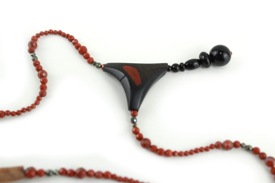Choker design en bois exotiques & Jaspe rouge, avec pour pendentif une vrille en bois d'ébène noir, attachée par un cercle de petite perle de pyrité bronze doré scintillant, reliée à une pièce balancelle courbée en bois d'olivier crème foncé boisé, et un fil de perles rouge brique ponctués de deux de pyrite partent des deux côté de la pièce jusqu'au fermoir puzzle aimanté de forme triangulaire, avec un pendant de perles noires en fermeture