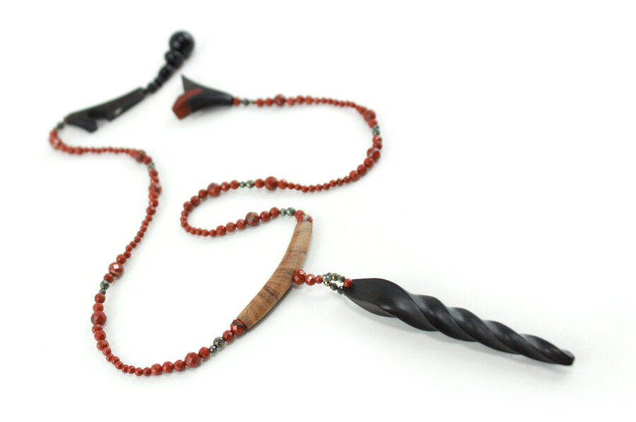 Choker design en bois exotiques & Jaspe rouge, avec pour pendentif une vrille en bois d'ébène noir, attachée par un cercle de petite perle de pyrité bronze doré scintillant, reliée à une pièce balancelle courbée en bois d'olivier crème foncé boisé, et un fil de perles rouge brique ponctués de deux de pyrite partent des deux côté de la pièce jusqu'au fermoir puzzle aimanté de forme triangulaire, avec un pendant de perles noires en fermeture