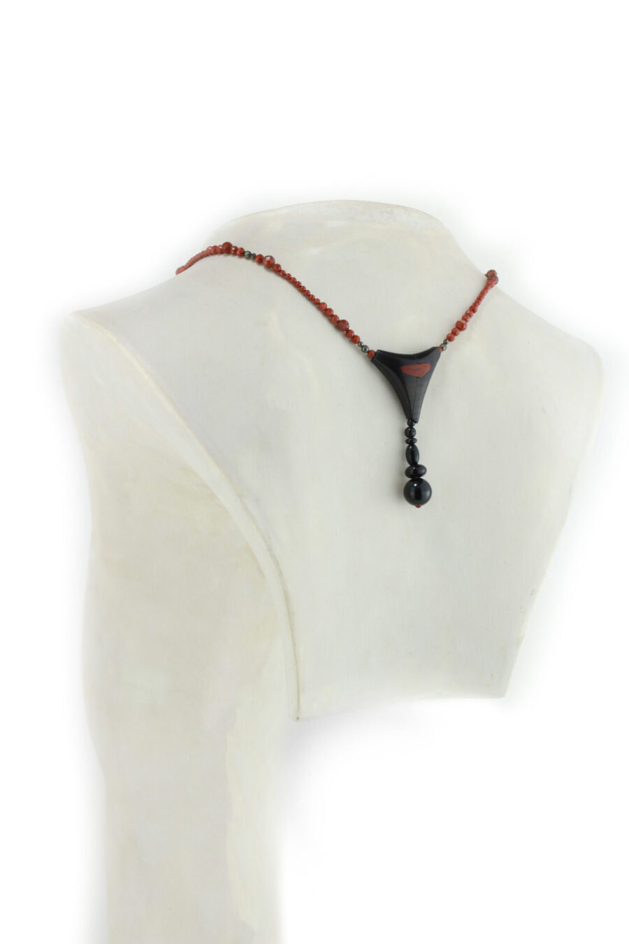 Choker design en bois exotiques & Jaspe rouge, avec pour pendentif une vrille en bois d'ébène noir, attachée par un cercle de petite perle de pyrité bronze doré scintillant, reliée à une pièce balancelle courbée en bois d'olivier crème foncé boisé, et un fil de perles rouge brique ponctués de deux de pyrite partent des deux côté de la pièce jusqu'au fermoir puzzle aimanté de forme triangulaire, avec un pendant de perles noires en fermeture