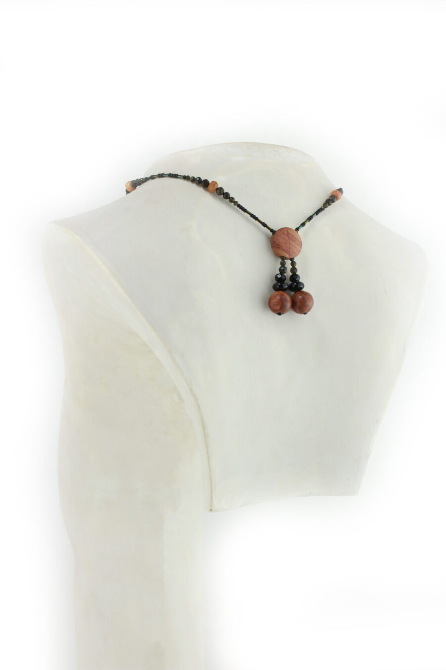 Collier croix primitive en bois : collier de perles noires, orange et vert métallisé fermé par un fermoir aimanté. Pendentif en forme de croix, bombée vers le haut, avec des lignes qui dessinent une croix sinueuse