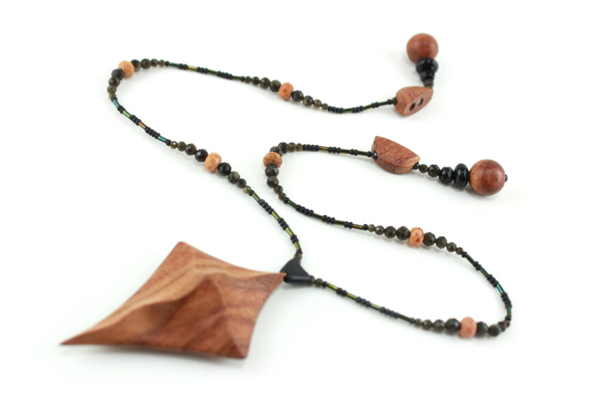 Collier croix primitive en bois : collier de perles noires, orange et vert métallisé fermé par un fermoir aimanté. Pendentif en forme de croix, bombée vers le haut, avec des lignes qui dessinent une croix sinueuse