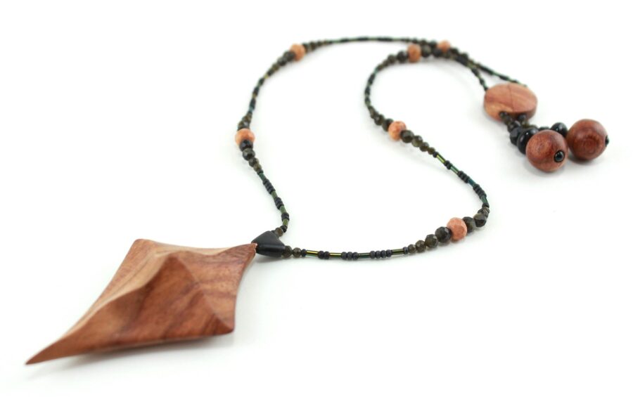 Collier croix primitive en bois : collier de perles noires, orange et vert métallisé fermé par un fermoir aimanté. Pendentif en forme de croix, bombée vers le haut, avec des lignes qui dessinent une croix sinueuse