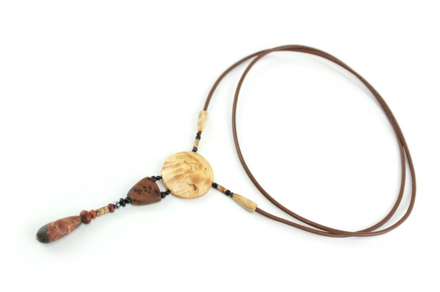 Collier sautoir en bois de Bouleau Madré sculpté & Picasso, avec en pendentif un disque crème boisé, une petite pièce en bois bombée marron tachetée de noir, une suite de perles et une pierre sculptée en forme d'olive vert tacheté d'orange. Cordon de cuir couleur camel relié aux pendant avec deux pièces de bois sculpté en forme d'olive