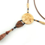 Collier sautoir en bois de Bouleau Madré sculpté & Picasso, avec en pendentif un disque crème boisé, une petite pièce en bois bombée marron tachetée de noir, une suite de perles et une pierre sculptée en forme d'olive vert tacheté d'orange. Cordon de cuir couleur camel relié aux pendant avec deux pièces de bois sculpté en forme d'olive