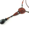 Sautoir en bois exotique et pierre d'Obsidienne, avec en pendentif un disque orange foncé zébré de noir, une petite pièce en bois bombée orange foncé, une suite de perles et une pierre sculptée en forme de goutte vert kaki. Cordon de cuir marron foncé relié aux pendant avec deux pièces de bois sculpté en forme d'olive