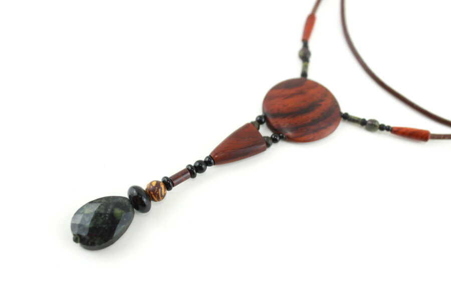 Sautoir en bois exotique et pierre d'Obsidienne, avec en pendentif un disque orange foncé zébré de noir, une petite pièce en bois bombée orange foncé, une suite de perles et une pierre sculptée en forme de goutte vert kaki. Cordon de cuir marron foncé relié aux pendant avec deux pièces de bois sculpté en forme d'olive