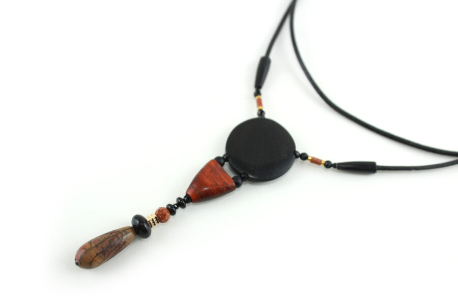 Collier totem en bois exotiques et Jaspe Picasso, avec en pendentif un disque noir ébène, une petite pièce en bois bombée orange foncé, une suite de perles et une pierre sculptée en forme d'olive vert tacheté d'orange. Cordon de cuir noir relié aux pendant avec deux pièces de bois sculpté en forme d'olive