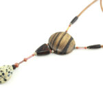 Sautoir magnétique en Ziricote & Jaspe dalmatien, avec en pendentif un disque noir grisé zébré, une petite pièce en bois bombée marron foncé, une suite de perles et une pierre sculptée en forme de goutte blanche tachetée de noir. Cordon de cuir orange doré relié aux pendant avec deux pièces de bois sculpté en forme d'olive