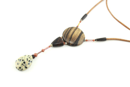 Sautoir magnétique en Ziricote & Jaspe dalmatien, avec en pendentif un disque noir grisé zébré, une petite pièce en bois bombée marron foncé, une suite de perles et une pierre sculptée en forme de goutte blanche tachetée de noir. Cordon de cuir orange doré relié aux pendant avec deux pièces de bois sculpté en forme d'olive